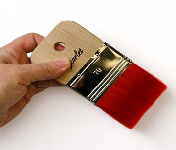 A hand is shown holding a wide rectangular shaped paint brush with scarlet red bristles against a white background. 