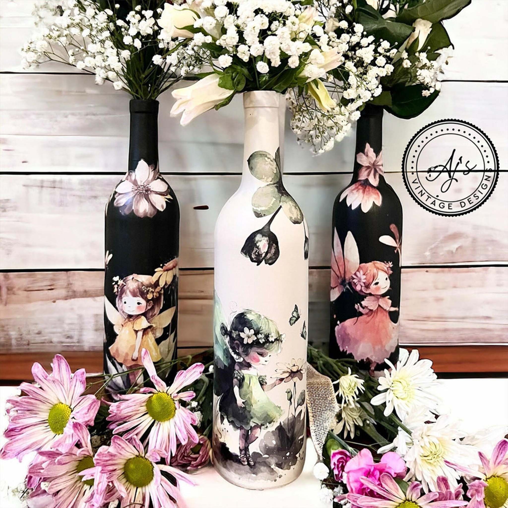 Three wine bottles refurbished by Aj's Vintage Designs feature Belles & Whistles' Fairyland small transfers on them. Two bottles are painted black and one is white.