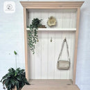 Wooden wardrobe with shelves, plants, and a handbag against a white brick wall.