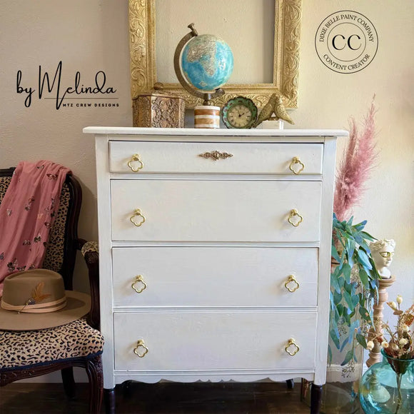White dresser with gold knobs in a room with decor elements like a chair, plant, and framed picture.