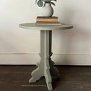 Round side table with a vase and books on a wooden floor against a white wall.