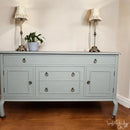 Light blue sideboard with two lamps and a plant on a wooden floor.
