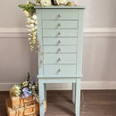 Light blue jewelry armoire with silver knobs against a neutral wall.