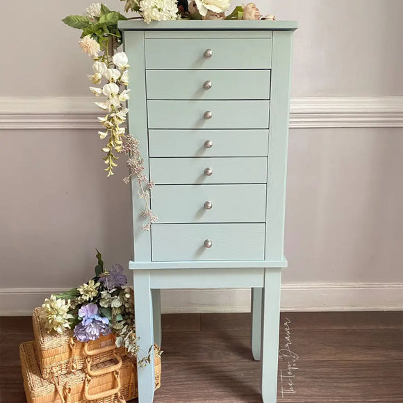 Light blue jewelry armoire with silver knobs against a neutral wall.