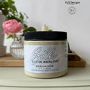 Jar of Silk All-in-One Mineral Paint with a black lid on a surface, with a blurred background featuring a pink flower and a plant.