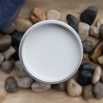 An arial view of an open container of Dixie Belle Paint Company's Glacier All-In-One Silk Paint is on a wood table with river rocks.
