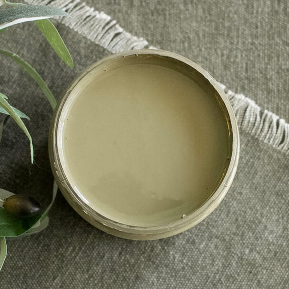 An arial view of an open container of Dixie Belle Paint Company's Hampton Olive All-In-One Silk Paint is on a grey material with white fringe and green foliage.