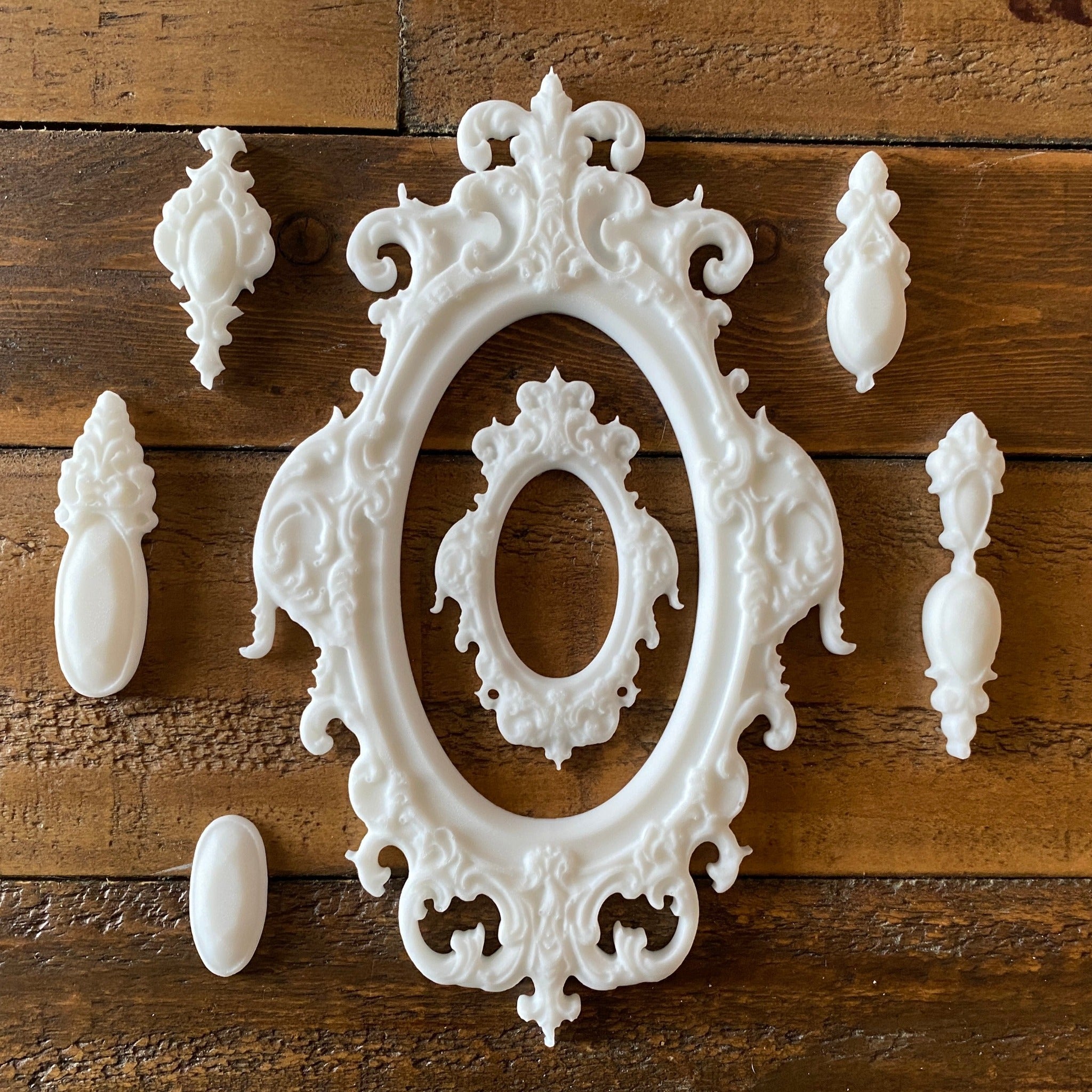 White silicone mould castings of 2 ornate oval frames and 5 ornate pendants are against a wood background.