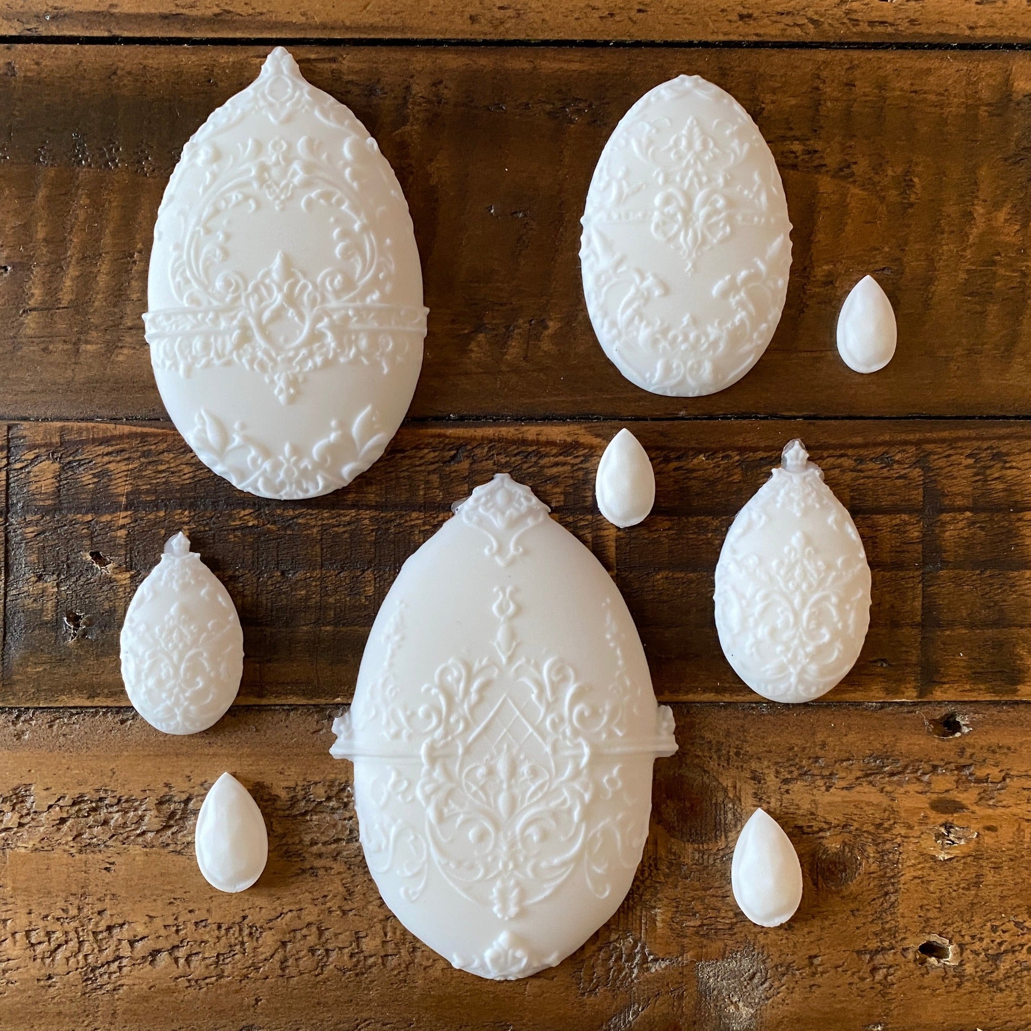White silicone mould castings of 5 varying sizes of ornately decorated Easter eggs and 4 small teardrop pendants are against a wood background.