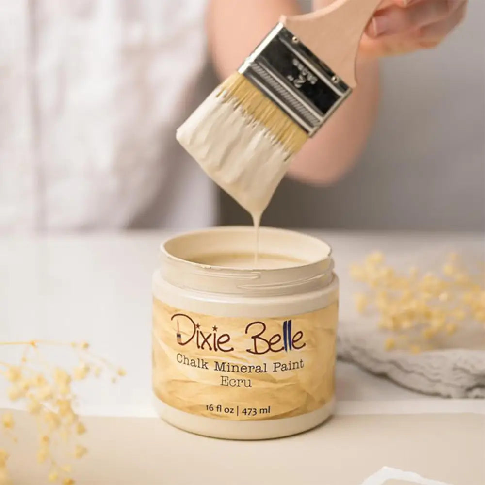 A paint brush with dripping paint is shown above an open container of Dixie Belle's Ecru Chalk Mineral Paint.