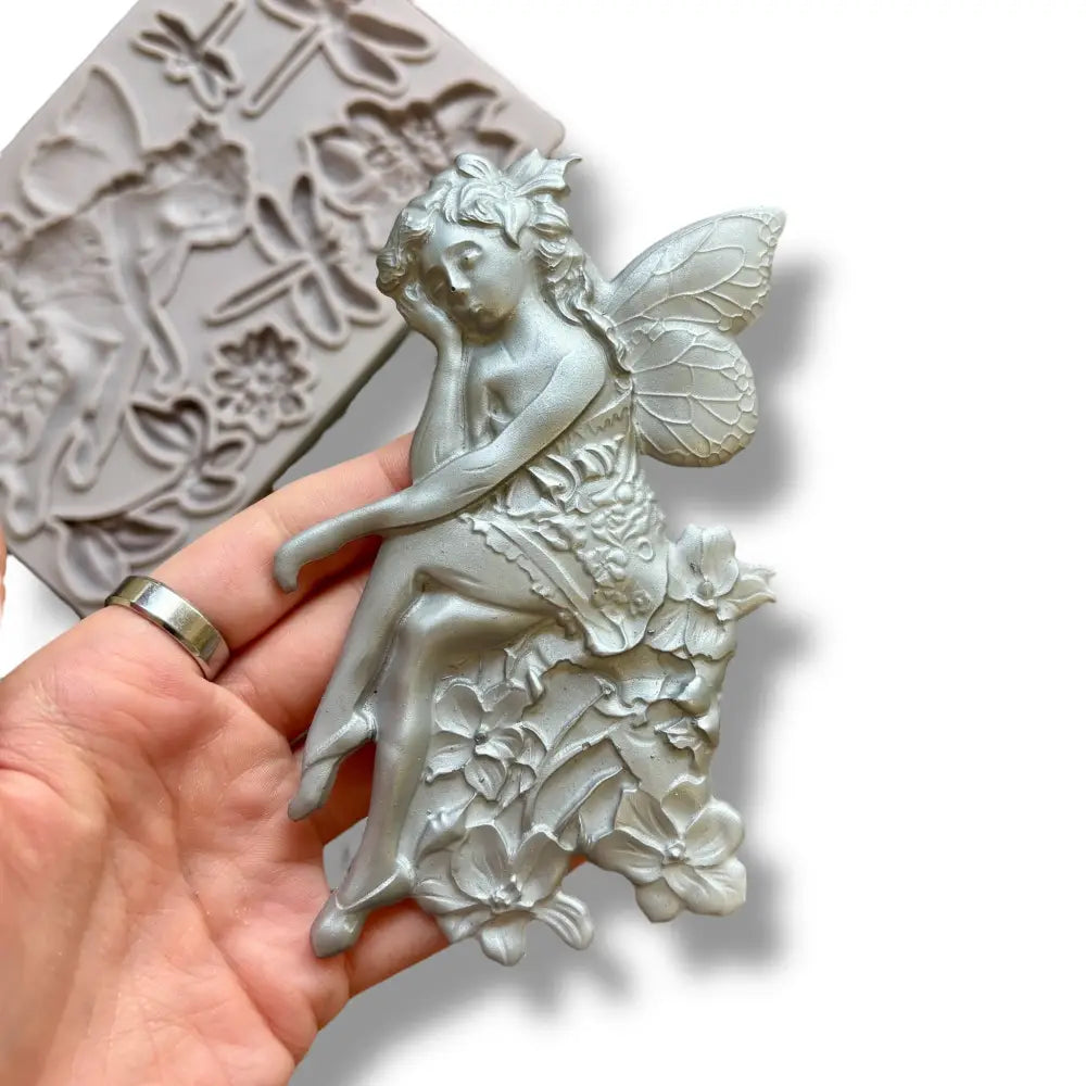 A light grey silicone mold featuring a fairy sitting on a cluster of flowers, 2 single flowers, leaves, and 3 dragonflies is against a white background. A hand is shown holding a silver colored casting of the fairy.