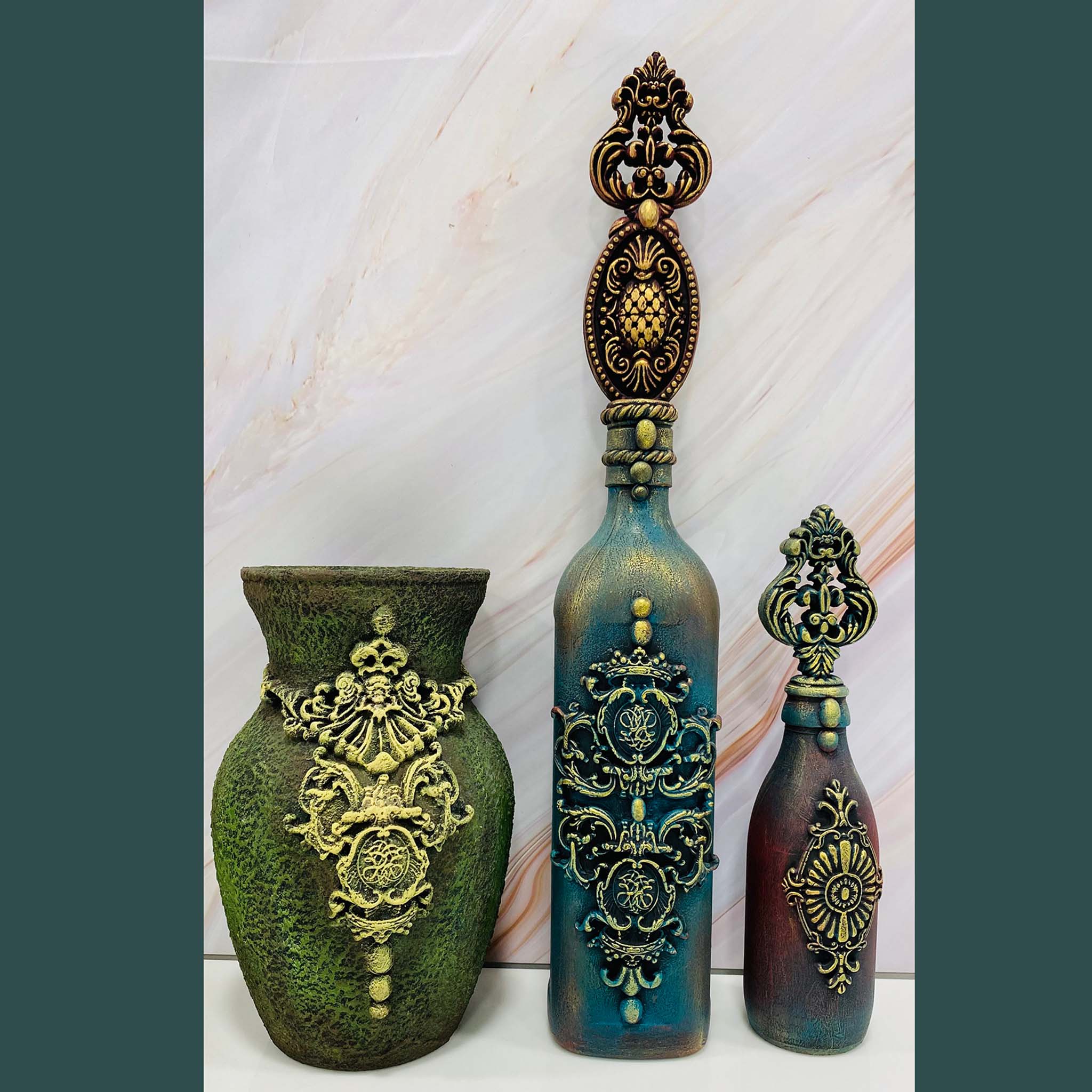 A green vase, and a purple and blue glass bottle feature Dadarkar Art by Get Inspired's Crowns of Bavaria silicone mold castings painted in gold on them.