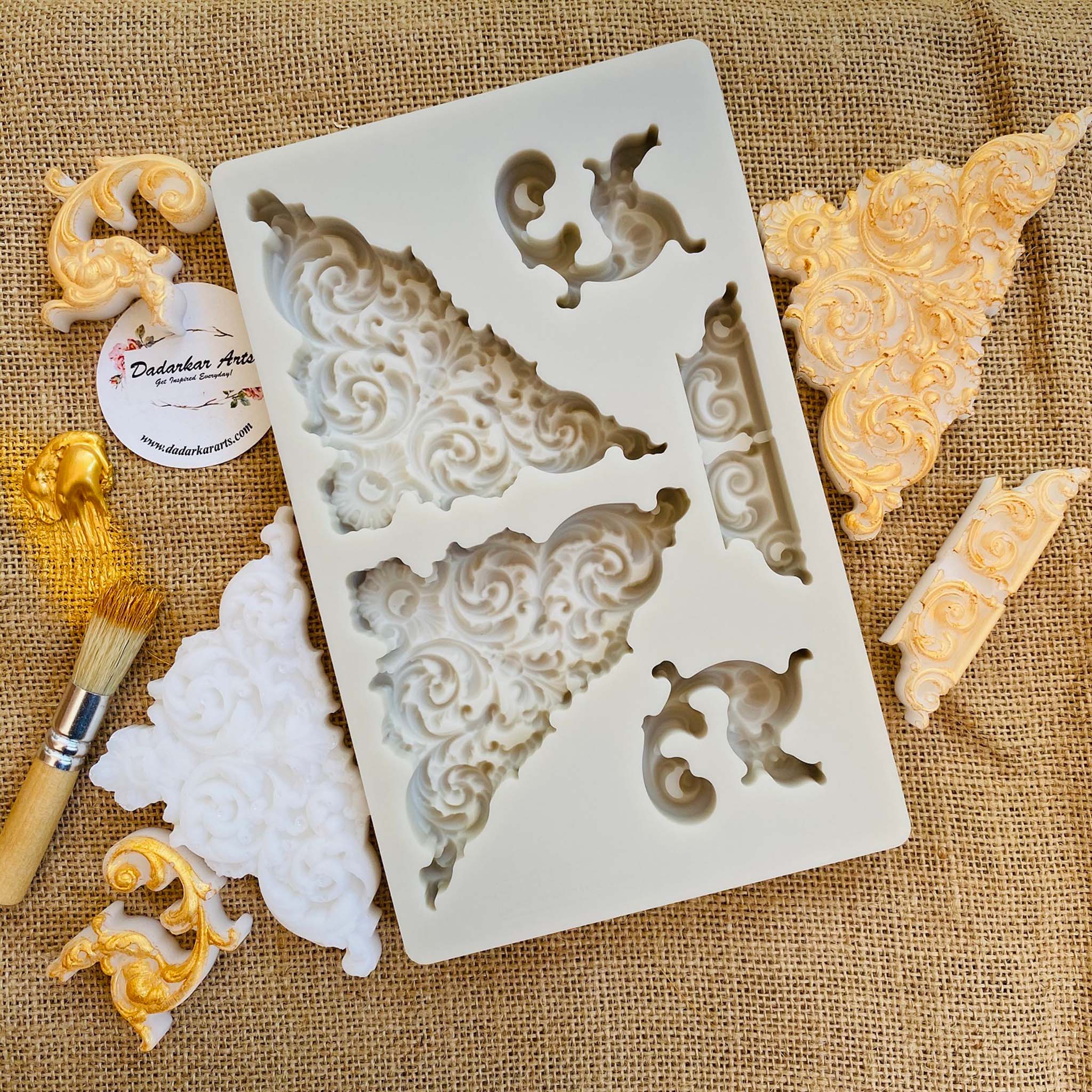 A light beige silicone mold and gold accented white resin castings of ornate corner and accent pieces are against a burlap background.