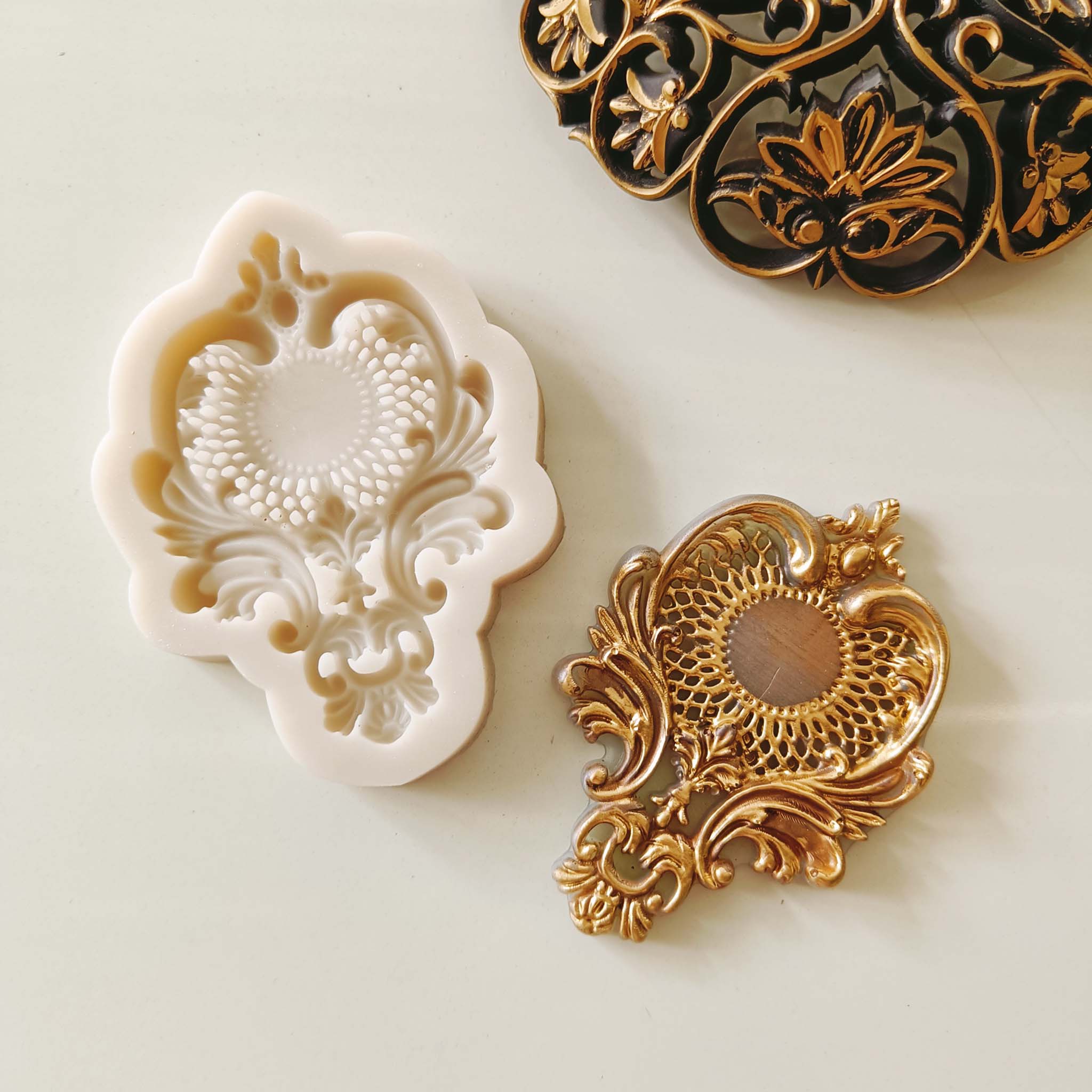 A light beige silicone mold and gold colored casting of a large chandelier style crown medallion are against a light beige background.