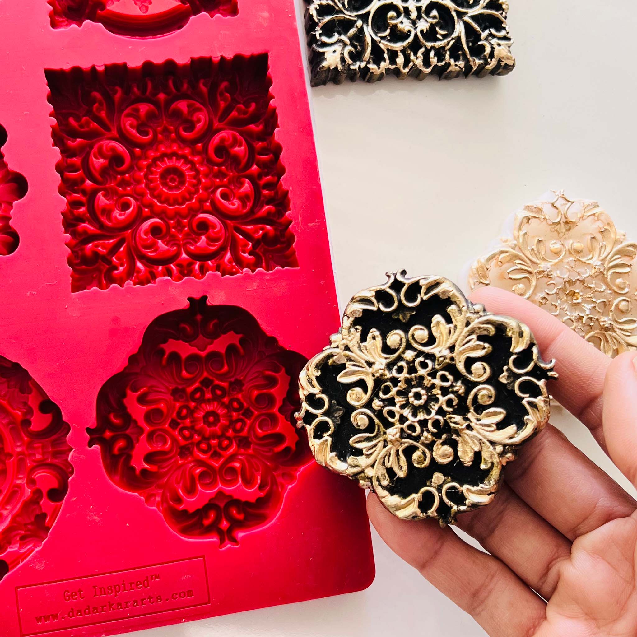 A hand is shown holding a dark gold colored casting against a red silicone mold of Dadarkar Art's Medallions Eminence. 