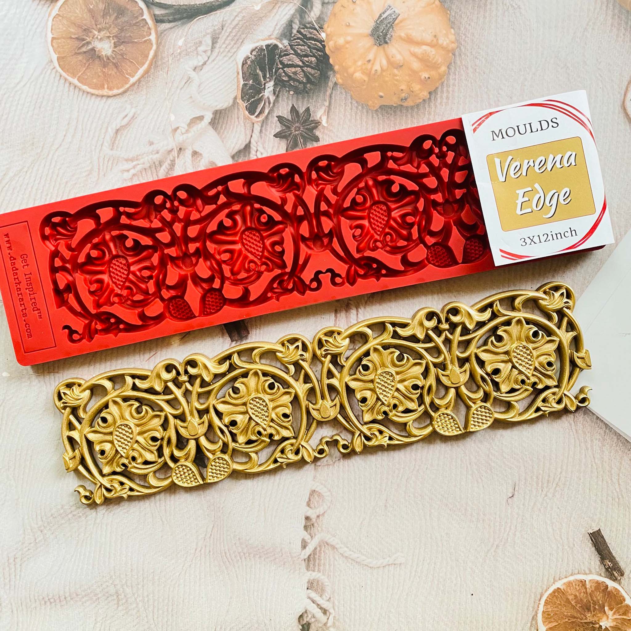 A red silicone mold and gold colored casting of Dadarkar Art's Verena Edge are against a light colored background with dried orange slices and a mini pumpkin.