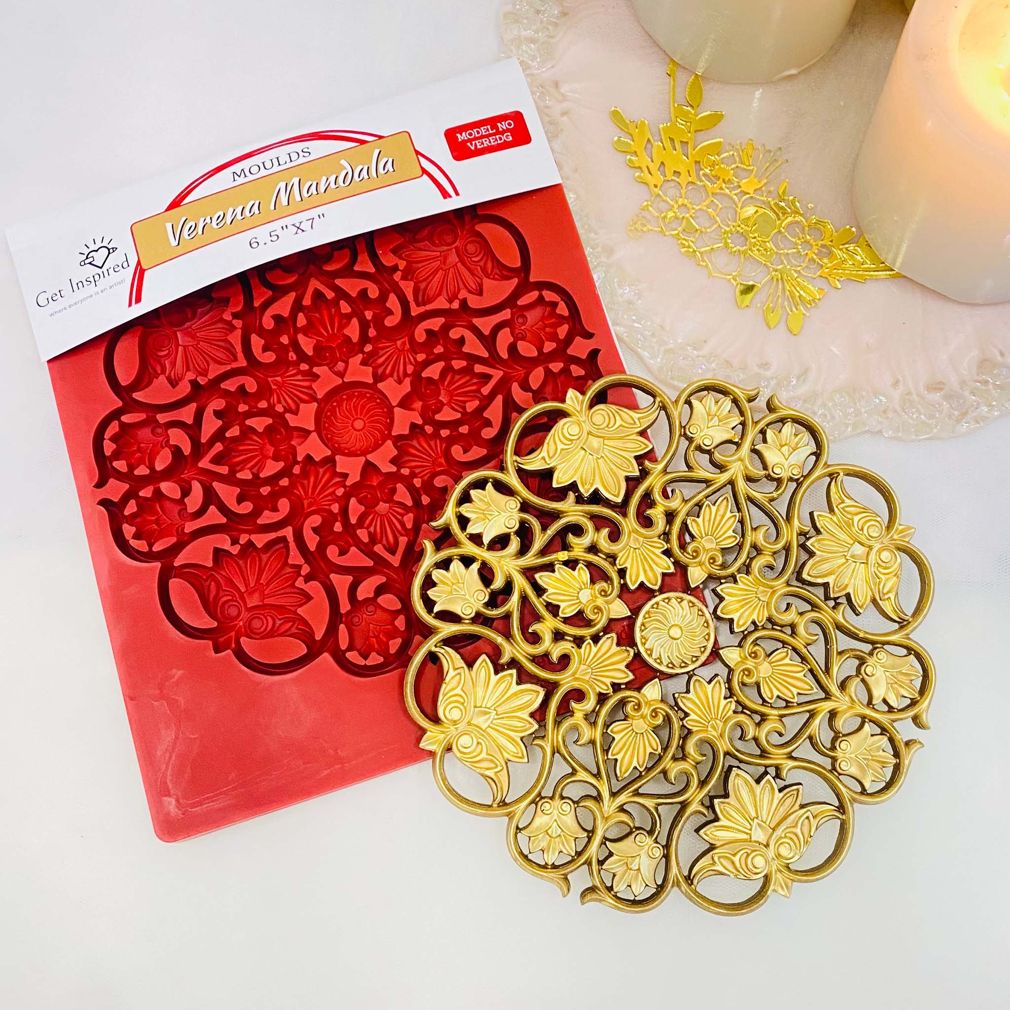 A gold colored casting and package of Get Inspired by Dadarkar Art's Verena Mandala silicone mold are on a light colored table with candles.