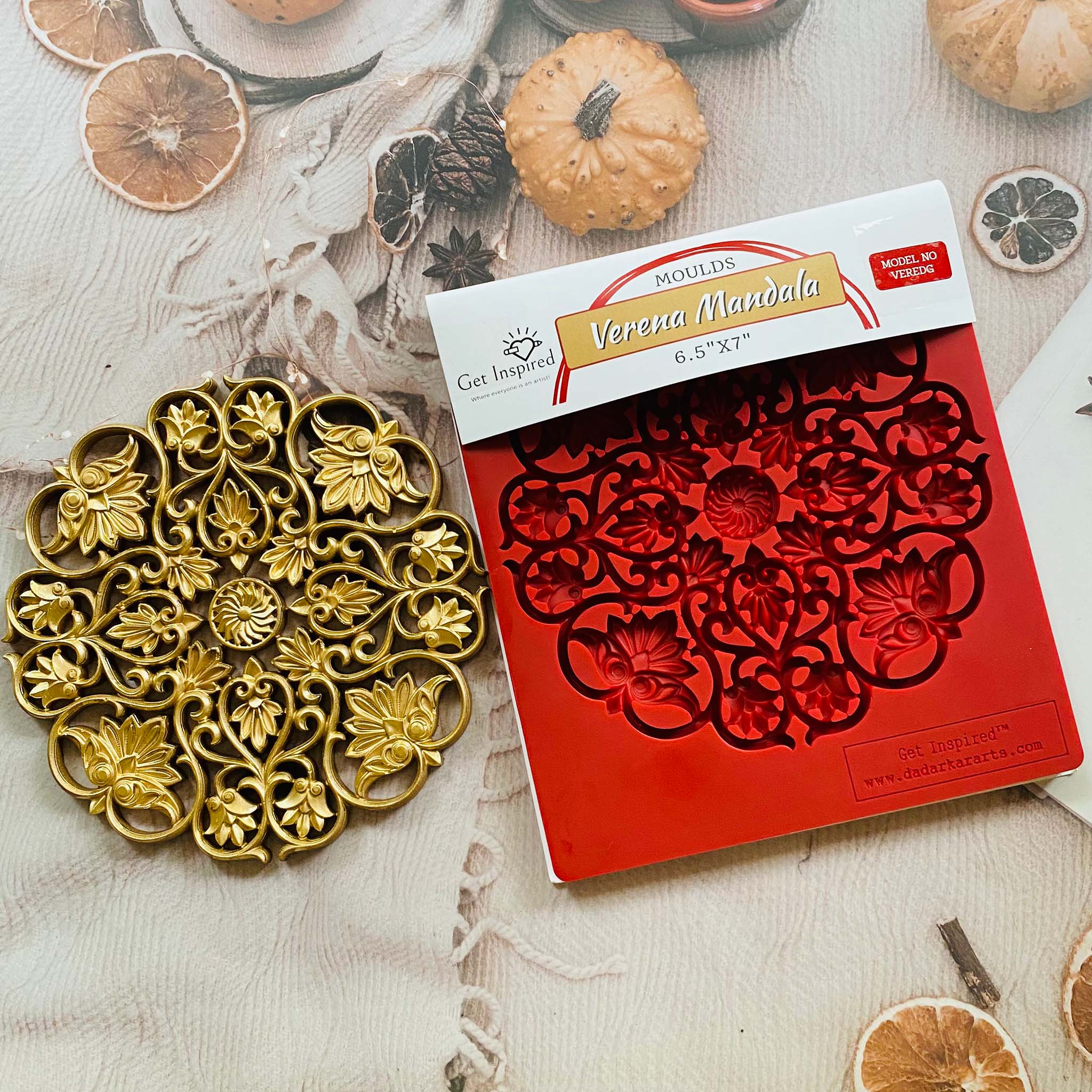 A gold colored casting and package of Get Inspired by Dadarkar Art's Verena Mandala silicone mold are on a table with dried orange slices and mini pumpkins.