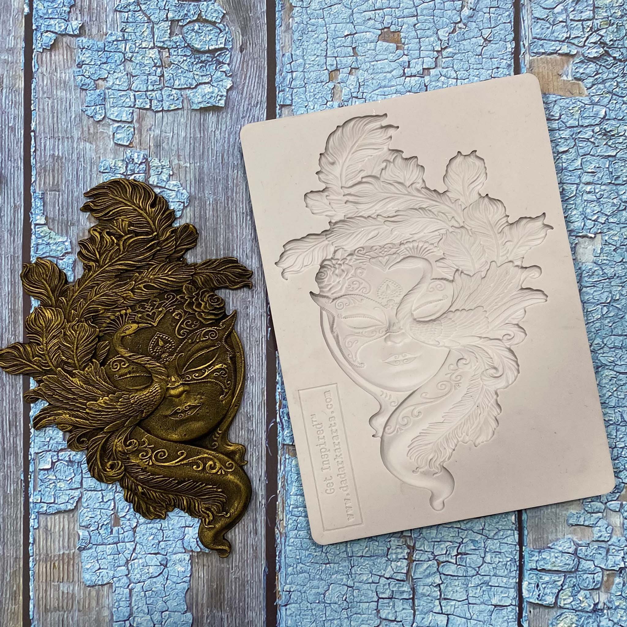 A light grey silicone mold and dark gold colored casting of a masquerade mask that features a peacock are against a weathered blue painted wood background.