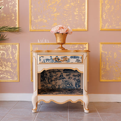 A vintage nightstand with 1 drawer and an open cubby shelf refurbished by Kacha features Kacha Furniture paint in the color Creme Brulee (neutral beige) and has gold accents with a blue toile rub-on transfer.