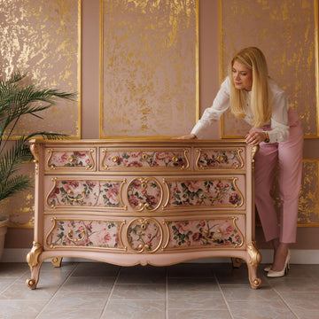 A vintage 5 drawer dresser refurbished by KACHA features KACHA Dusty Pink furniture paint with gold accents and floral details. KACHA herself is standing next to the dresser leaning slightly over it.