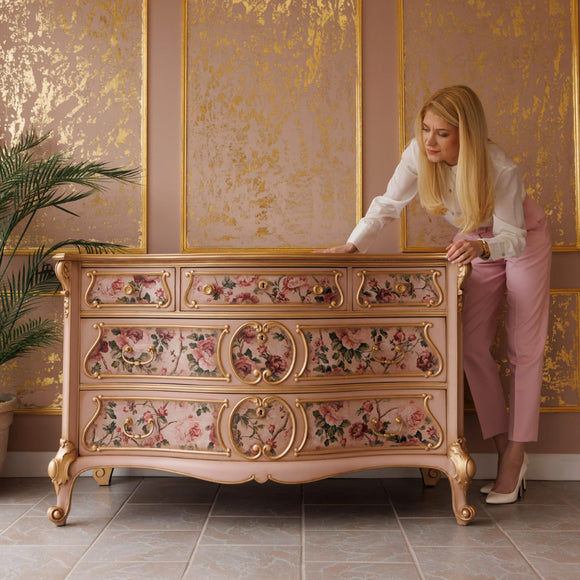 A vintage 5 drawer dresser refurbished by KACHA features KACHA Dusty Pink furniture paint with gold accents and floral details. KACHA herself is standing next to the dresser leaning slightly over it.