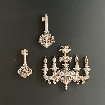 Silver colored silicone mold castings of a vintage chandelier and 2 vintage ornate keys are against a dark grey background.