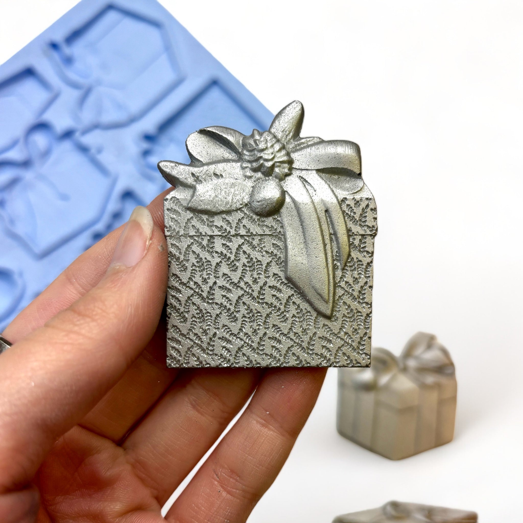 A blue silicone mold and silver colored castings of 5 beautifully wrapped packages, complete with a large bow and garland are against a white background. A hand is shown holding one of the gift castings.