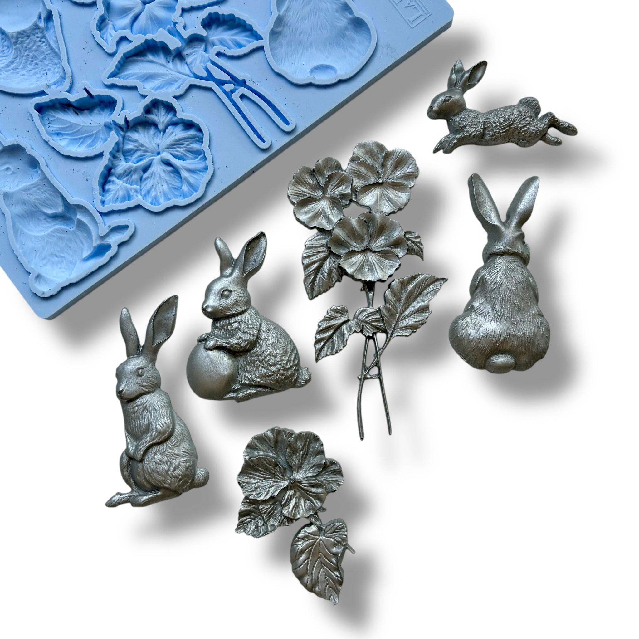 A blue silicone mold and silver colored castings featuring bunnies and a bouquet of flowers are against a white background.