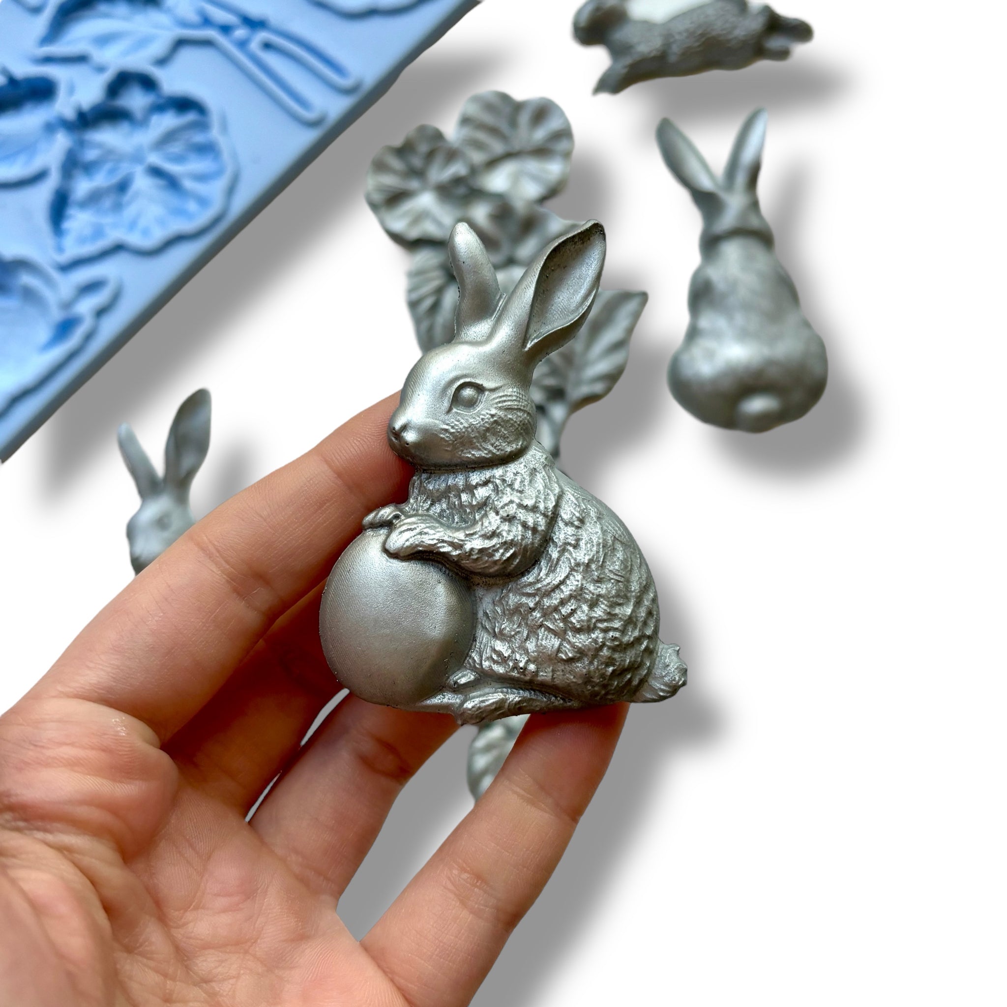 A blue silicone mold and silver colored castings featuring bunnies and a bouquet of flowers are against a white background. A hand is shown holding one of the bunny castings.