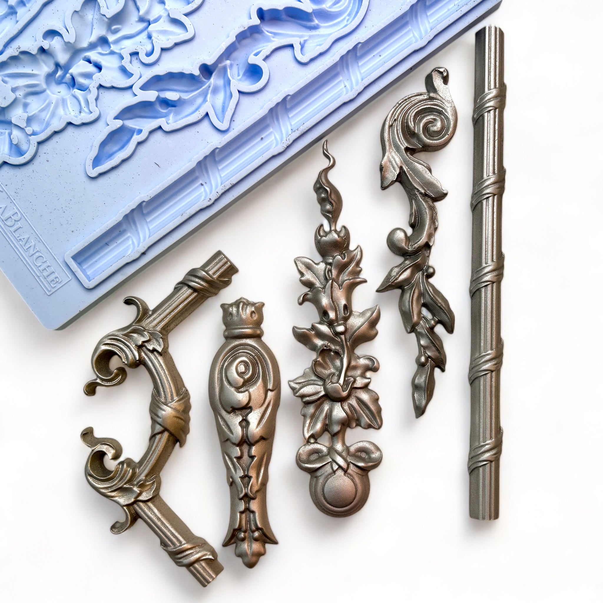 A blue silicone mold and silver colored castings of 5 ornate border trim and accent pieces are against a white background.