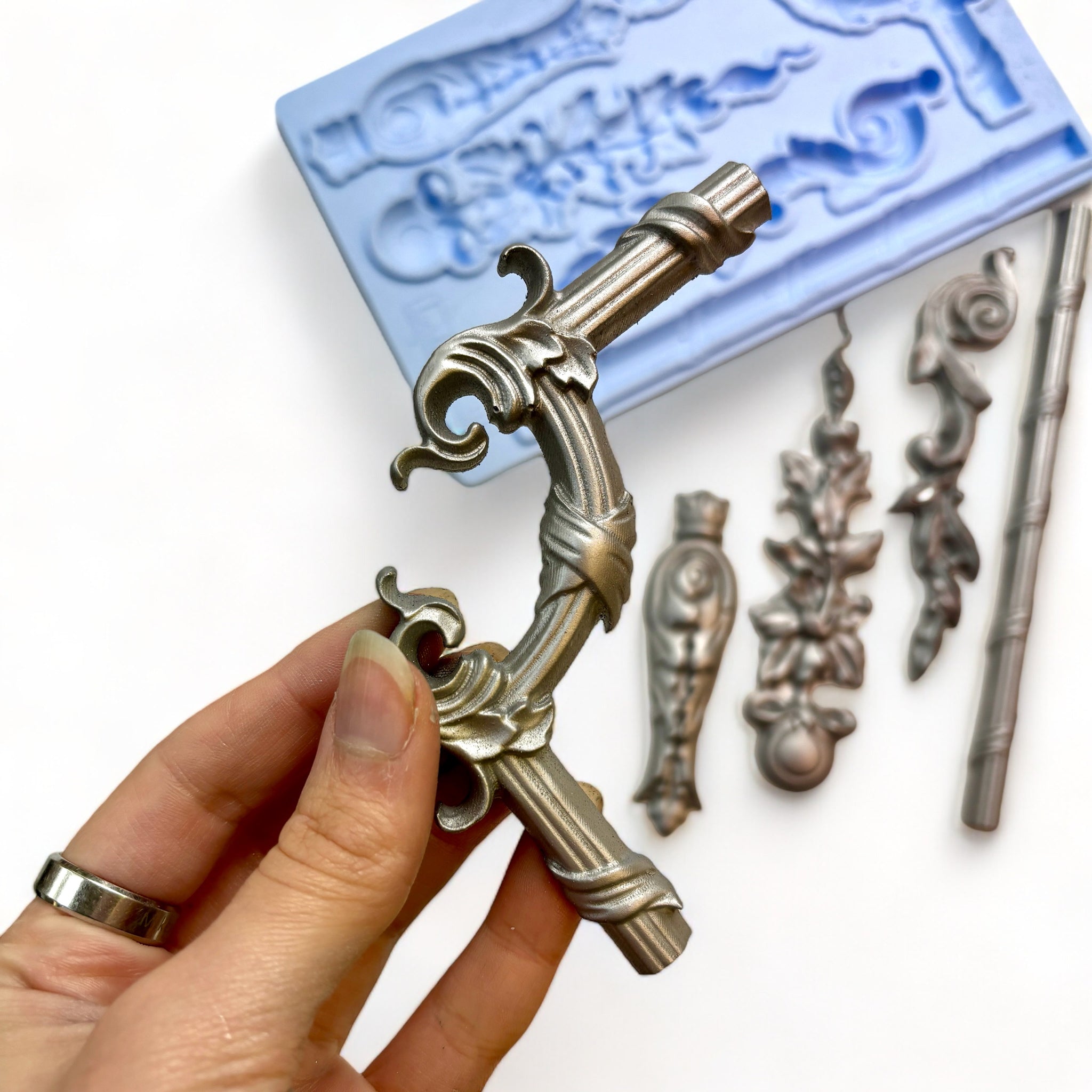 A blue silicone mold and silver colored castings of 5 ornate border trim and accent pieces are against a white background. A hand is shown holding one of the castings.