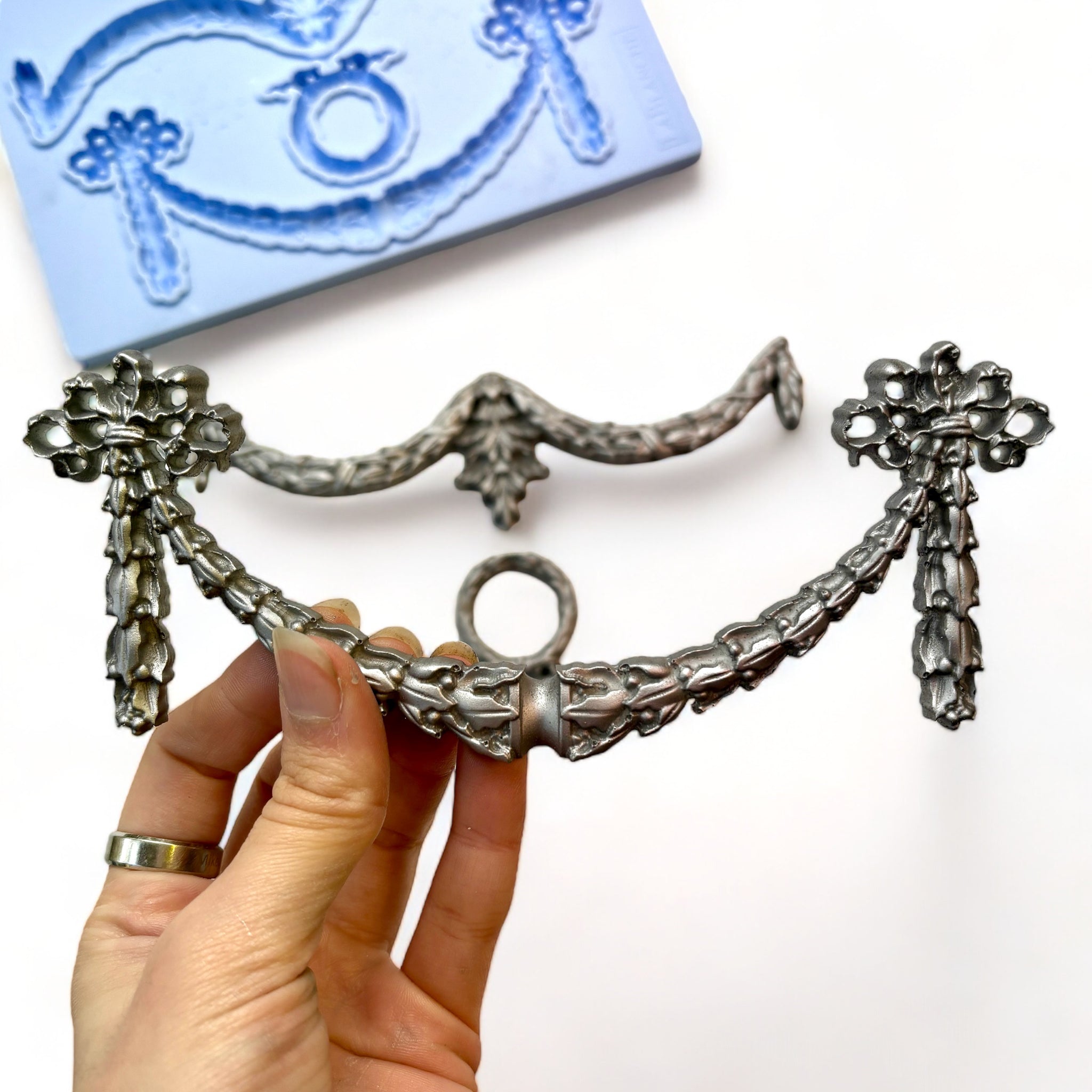 A blue silicone mold and silver colored castings of 2 draping garlands and a wreath are against a white background. A hand is shown holding one of the garland castings.