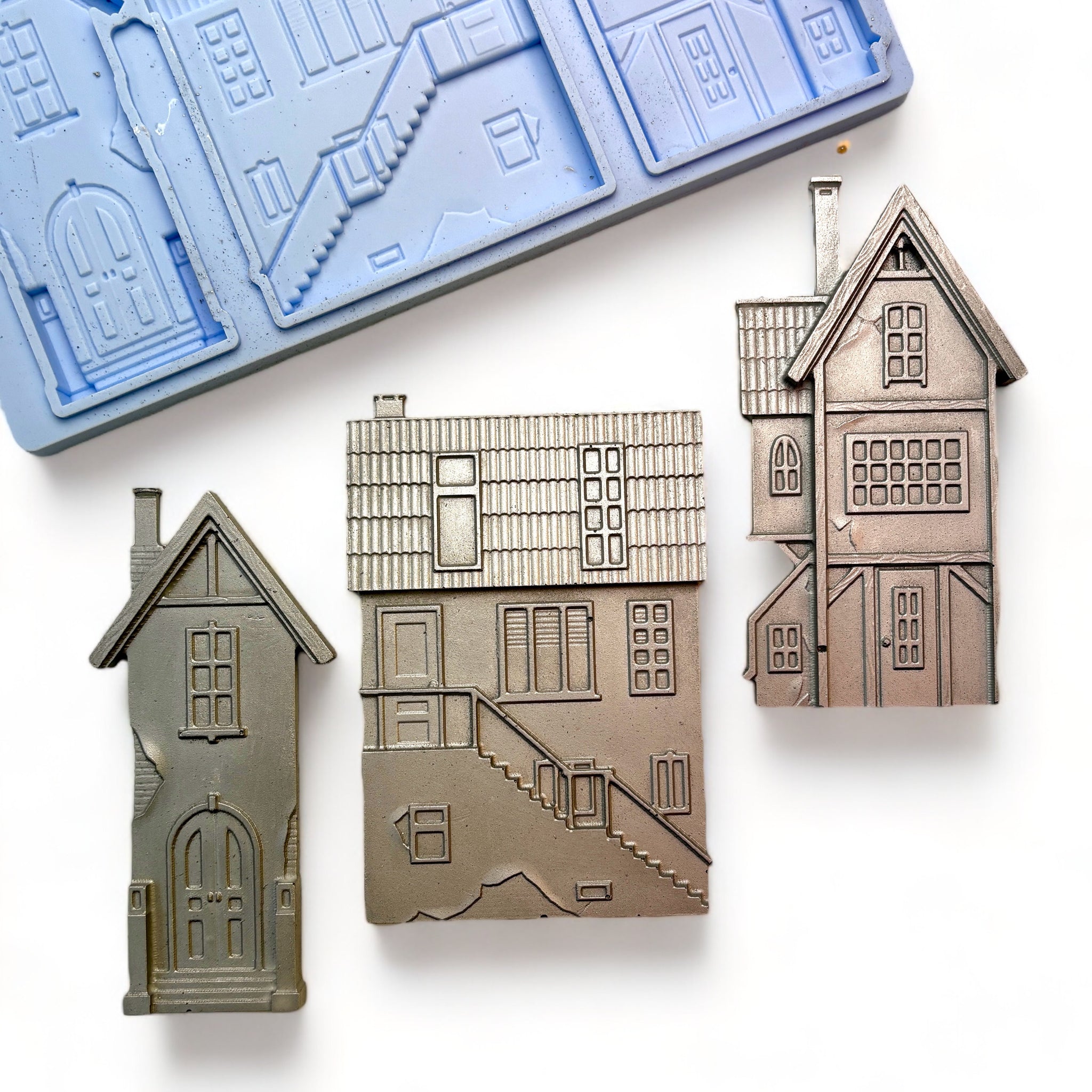 A blue silicone mold and silver colored castings of 3 unique 3-story homes are against a white background.