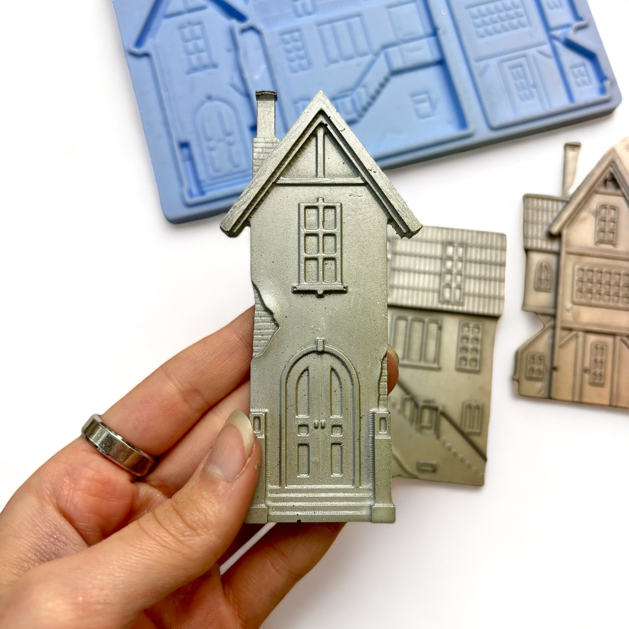 A blue silicone mold and silver colored castings of 3 unique 3-story homes are against a white background. A hand is shown holding one of the house castings.