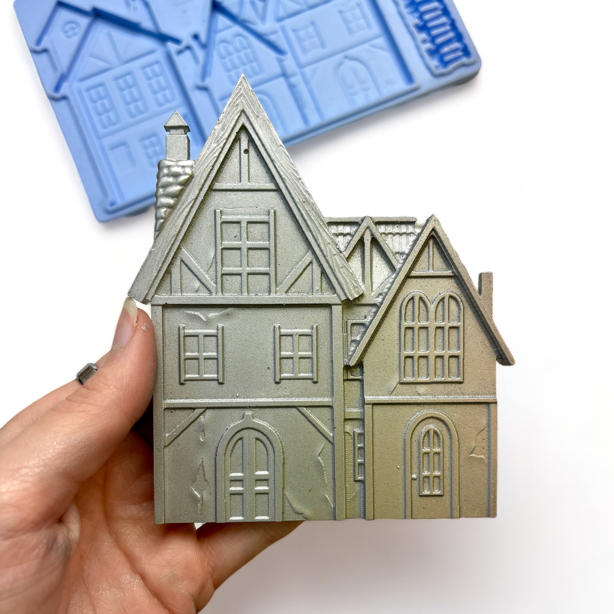 A blue silicone mold and silver colored castings of 2 homes, a picket fence, and a lamp post are against a white background. A hand is shown holding a house casting.