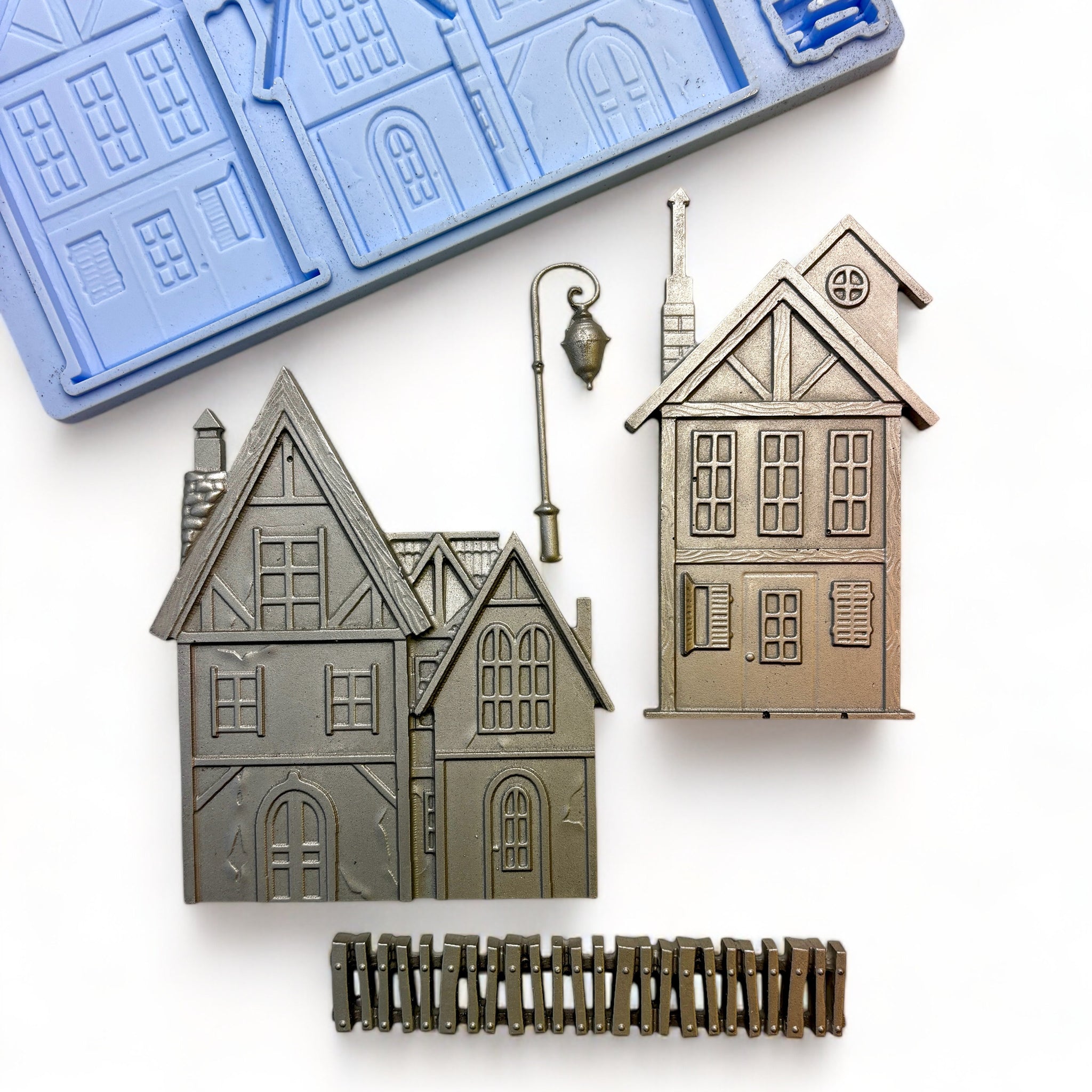 A blue silicone mold and silver colored castings of 2 homes, a picket fence, and a lamp post are against a white background.