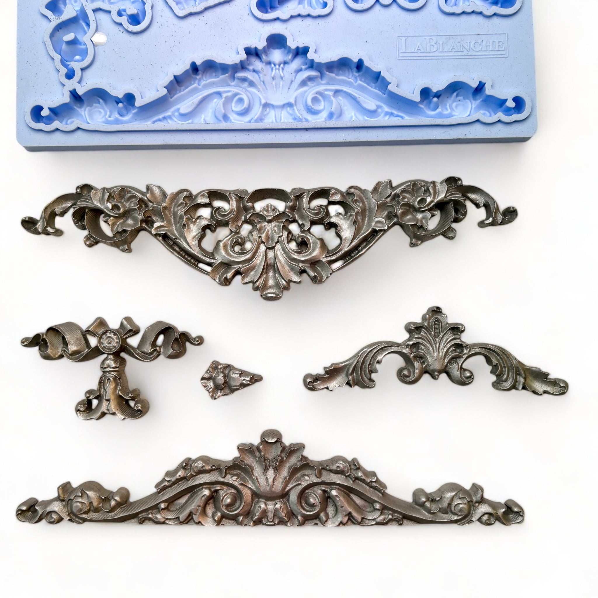 A blue silicone mold and silver  castings of 5 ornate scroll center accent pieces are against a white background.