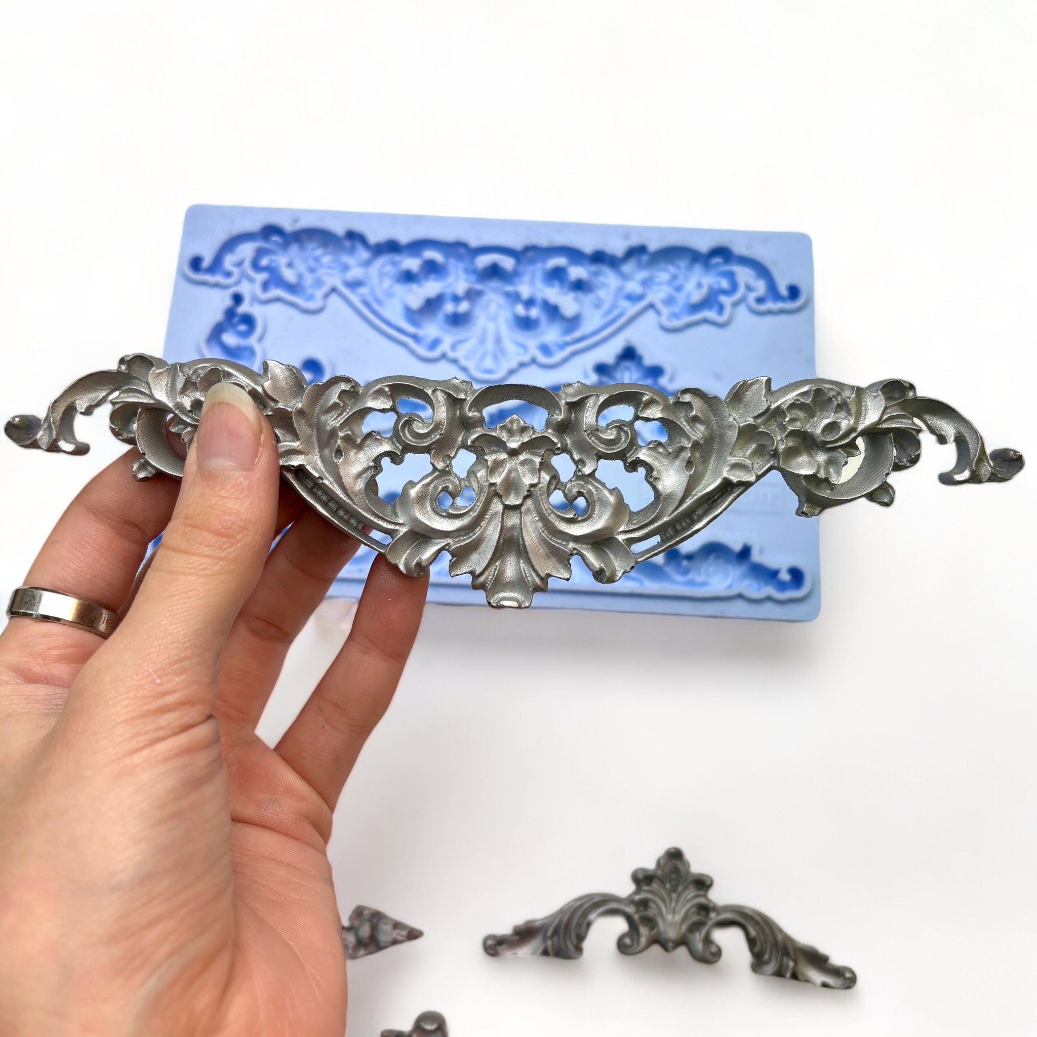 A blue silicone mold and silver  castings of 5 ornate scroll center accent pieces are against a white background. A hand is shown holding one of the castings.