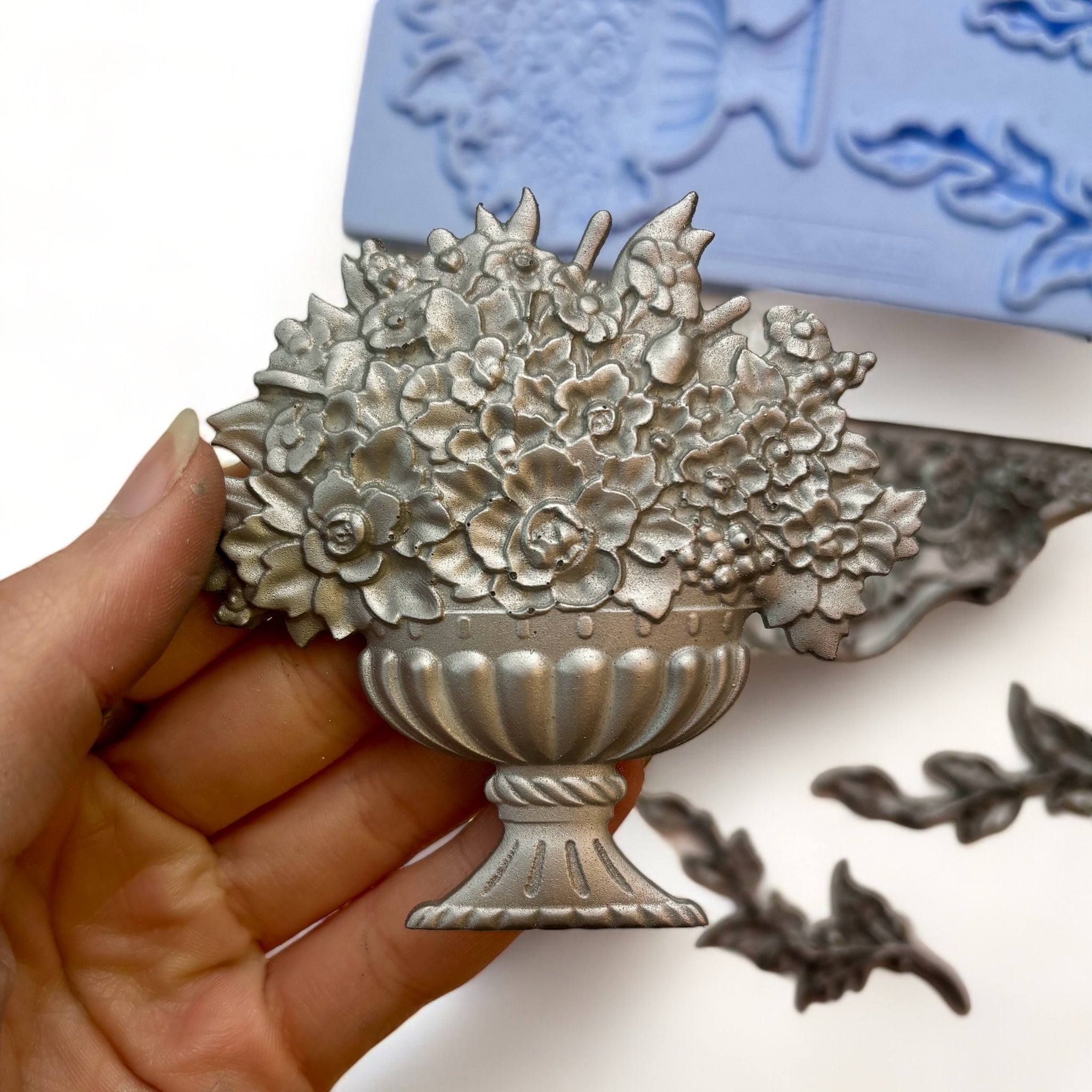 A blue silicone mold and silver colored castings of a planter pot with flowers, 2 vines, and a center accent piece are against a white background. A hand is shown holding the flower planter.