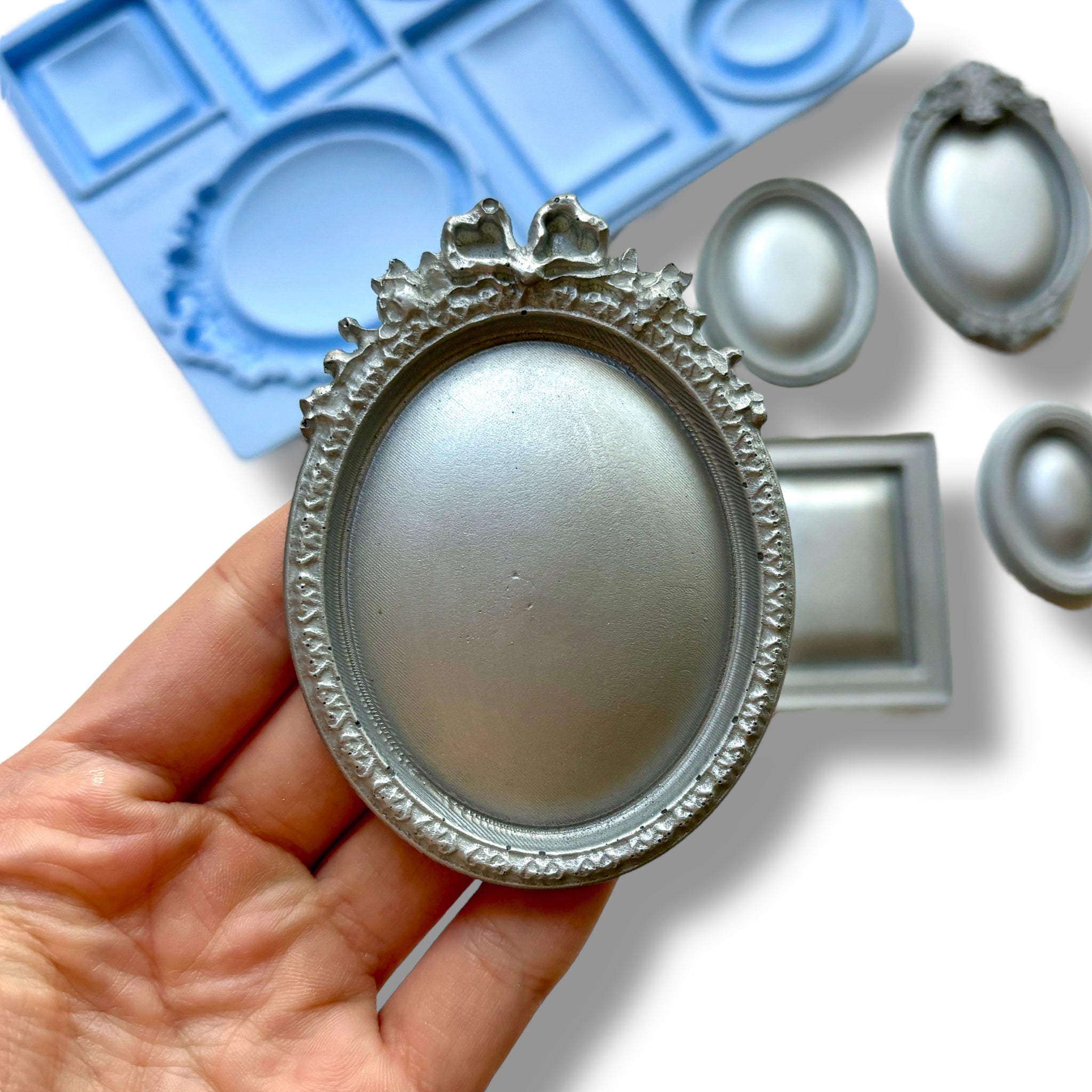 A blue silicone mold and silver colored castings featuring 7 small square, oval, and rectangular vintage photo frames are against a white background. A hand is shown holding one of the oval frames.