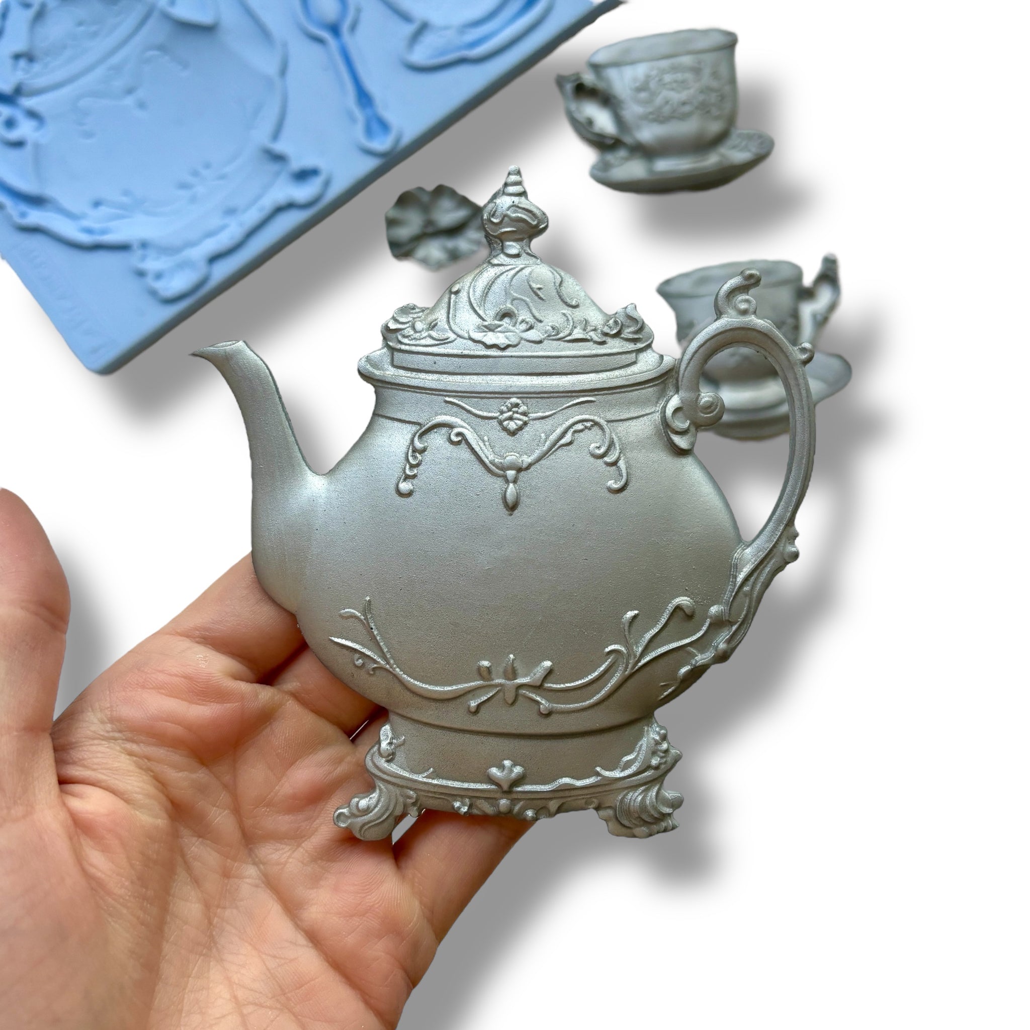 A blue silicone mold and silver colored castings of a tea set including a teapot, 2 cups, a spoon, and a flower are against a white background. A hand is shown holding the teapot casting.