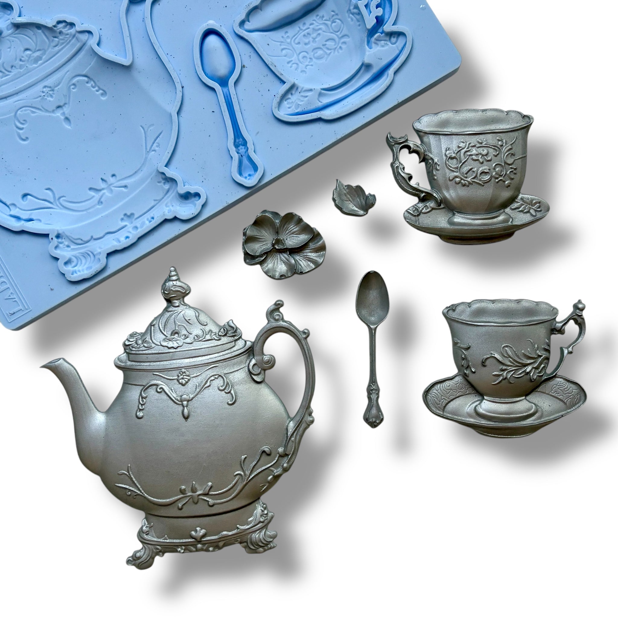 A blue silicone mold and silver colored castings of a tea set including a teapot, 2 cups, a spoon, and a flower are against a white background.