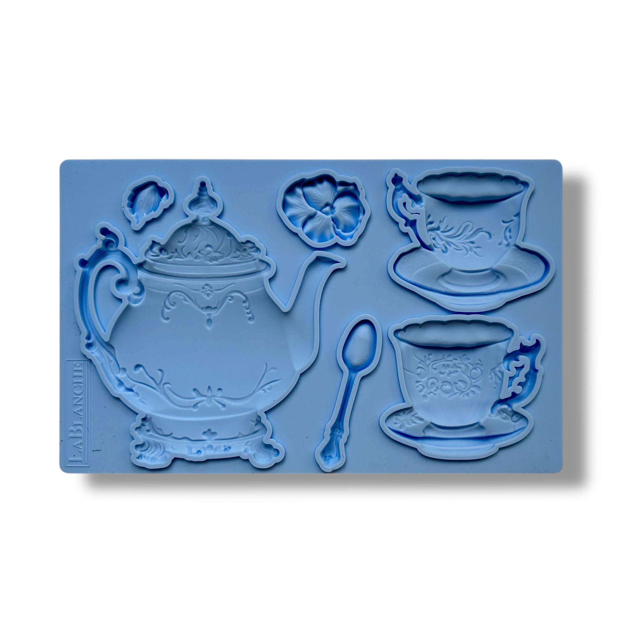 A blue silicone mold of a tea set including a teapot, 2 cups, a spoon, and a flower is against a white background.