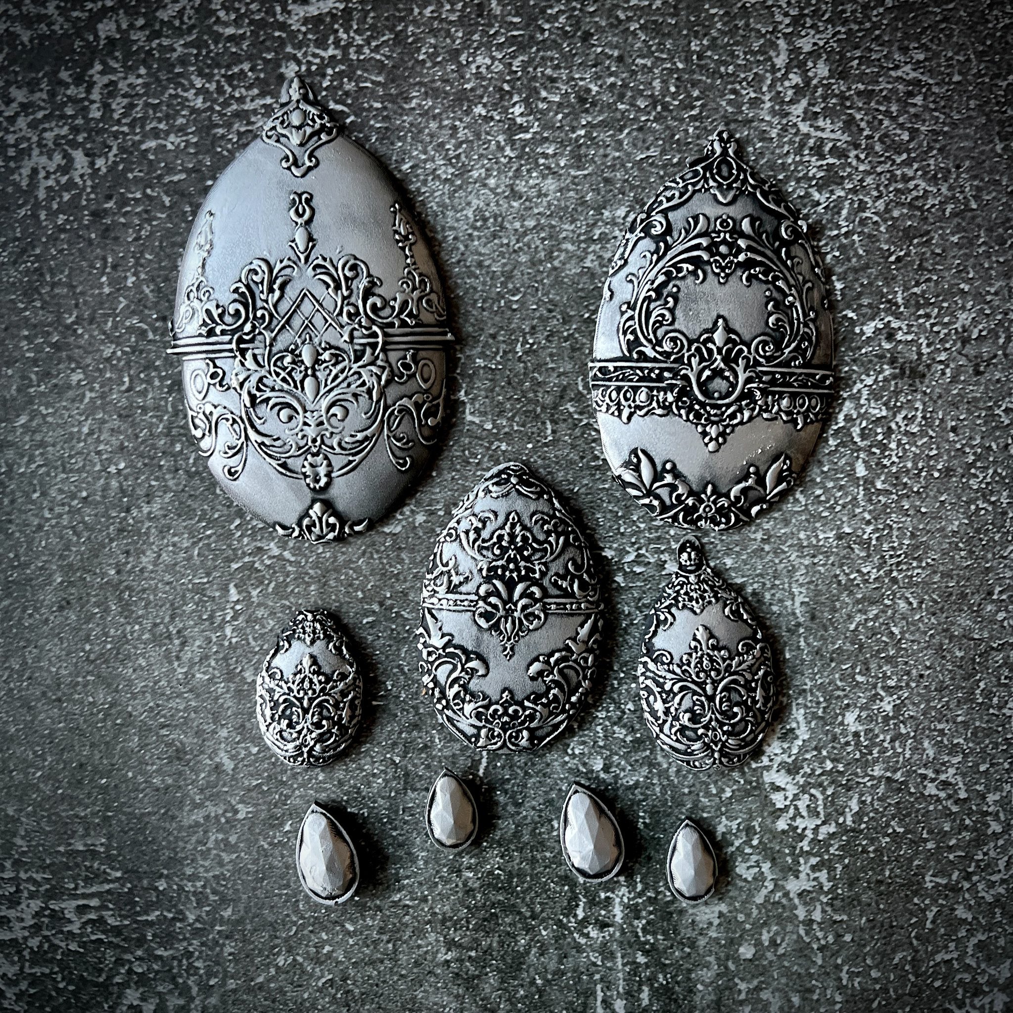 Silver silicone mould castings of 5 varying sizes of ornately decorated Easter eggs and 4 small teardrop pendants are against a textured grey background.