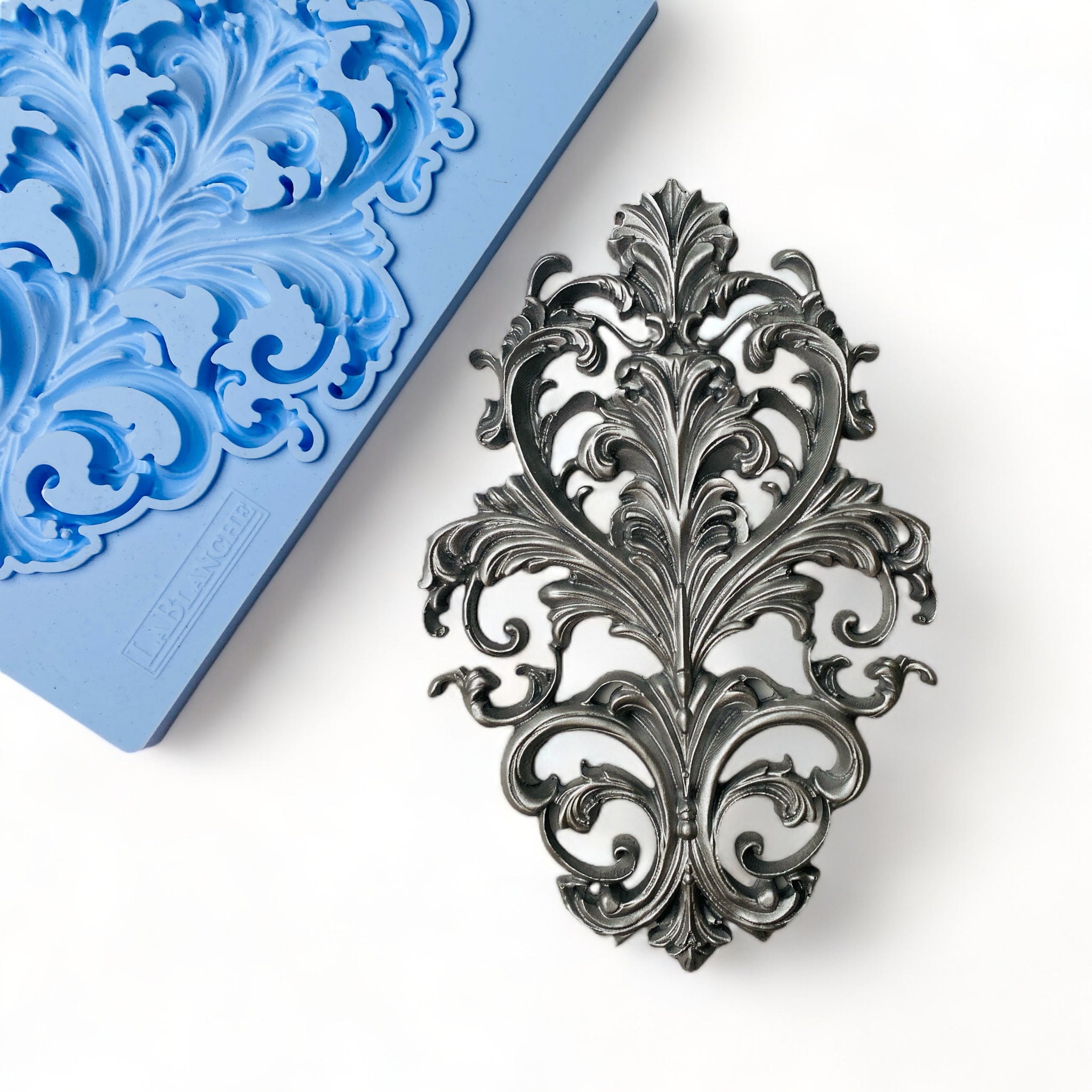 A blue silicone mold and silver colored casting of a baroque flourish emblem are against a white background.