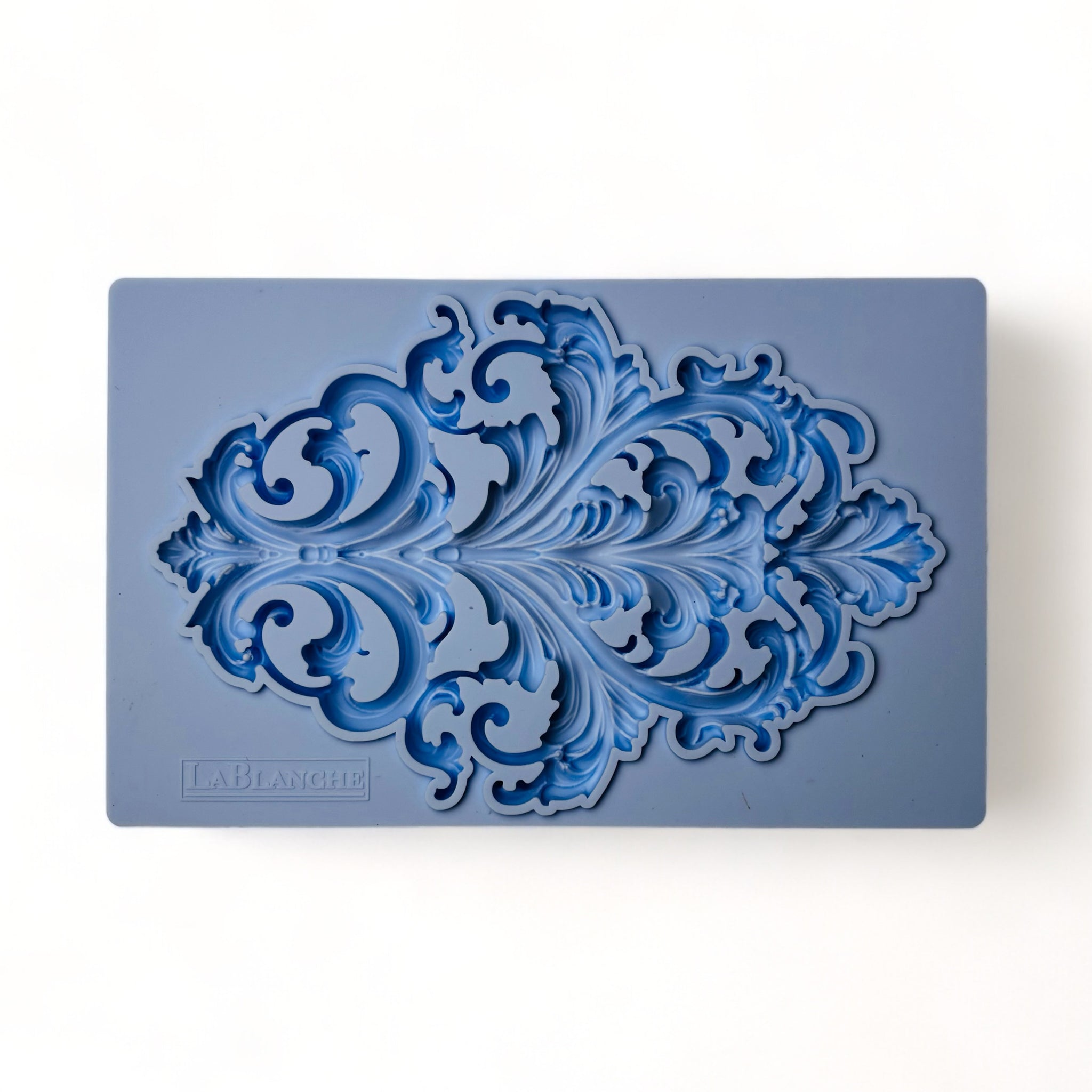 A blue silicone mold of a scrolling Baroque flourish emblem is against a white background.