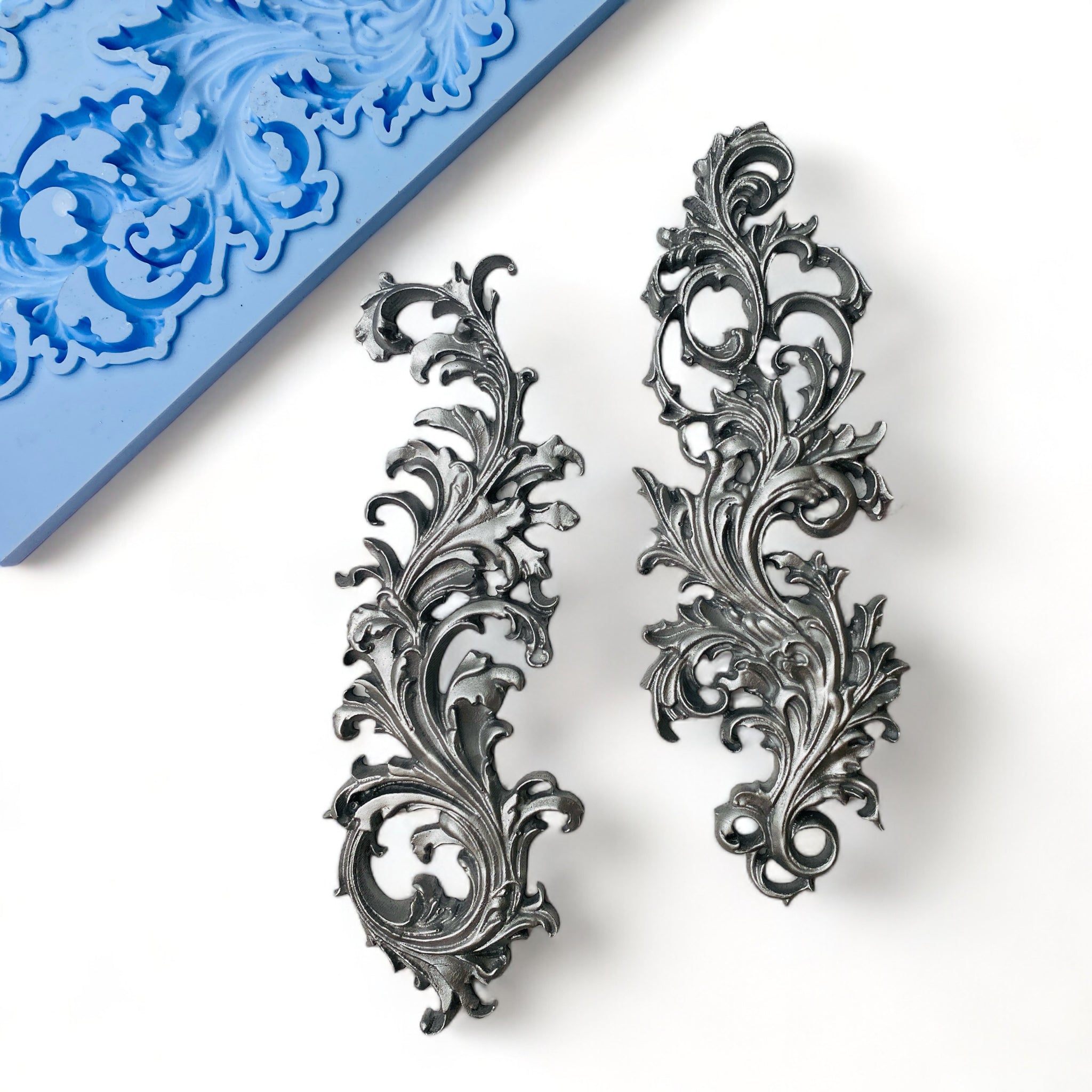 A blue silicone mold and silver colored casting of 2 leafy flourishes are against a white background.