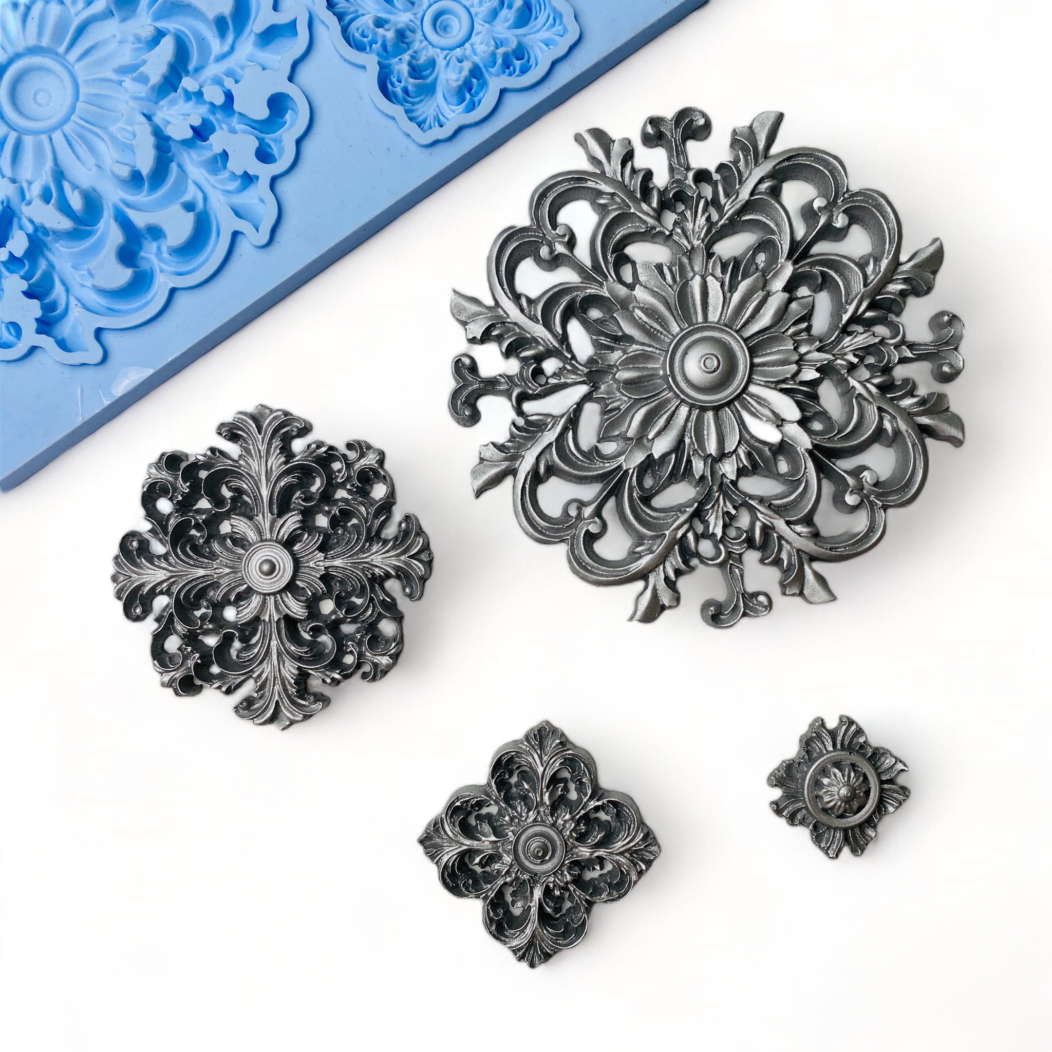 A blue silicone mold and silver colored castings that features 4 varying size round ornate medallions are against a white background.