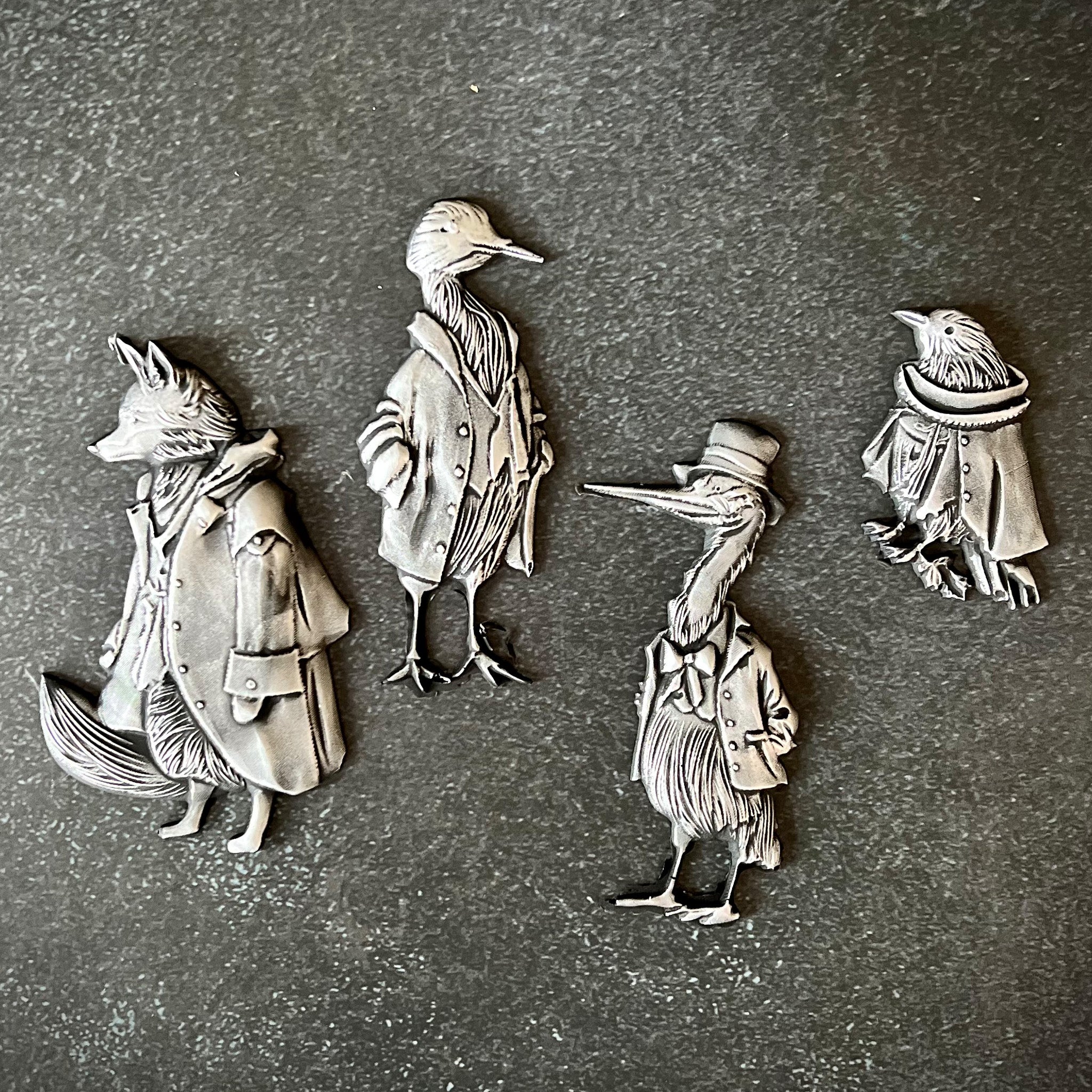 Silver colored castings of a fox and 3 birds all wearing vintage overcoats are against a grey background.